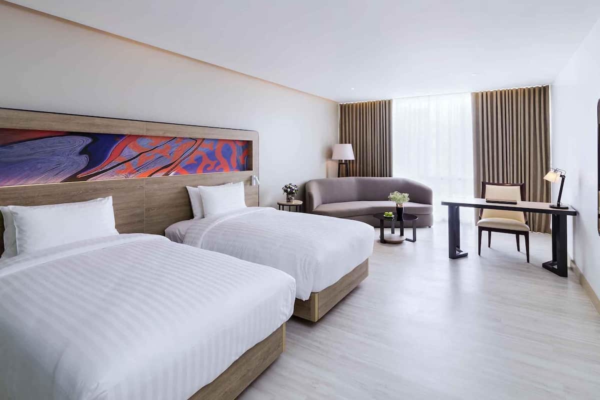 Quarto de hotel com duas camas, uma escrivaninha, uma cadeira, um abajur e um quadro grande de arte abstrata na parede.
