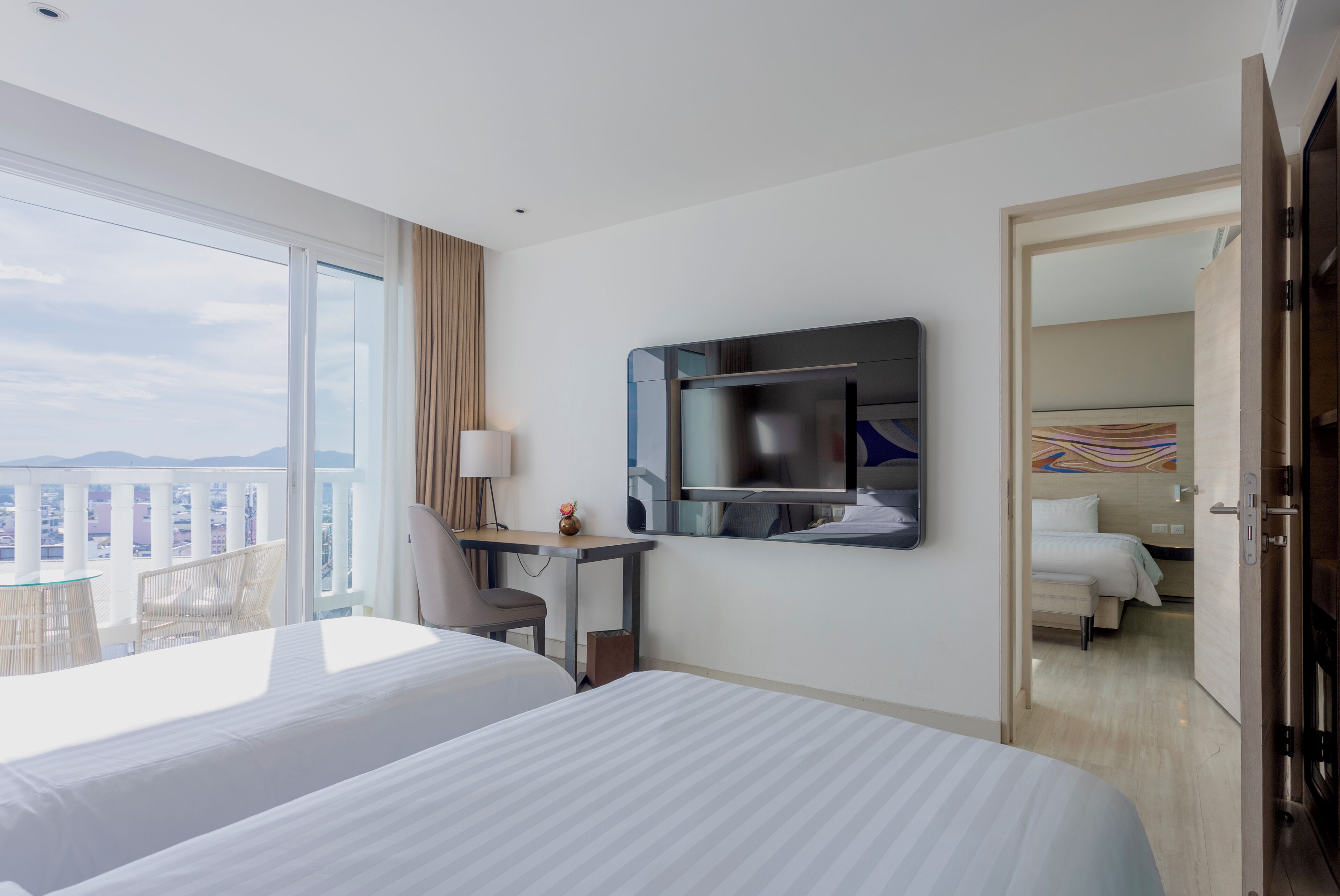 A hotel room with two beds, a desk, a chair, a TV, and a balcony with a view of the city.