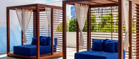 Dos camas tapizadas de azul con estructuras de madera y cortinas blancas, frente a una pared con un diseño de hojas azules.