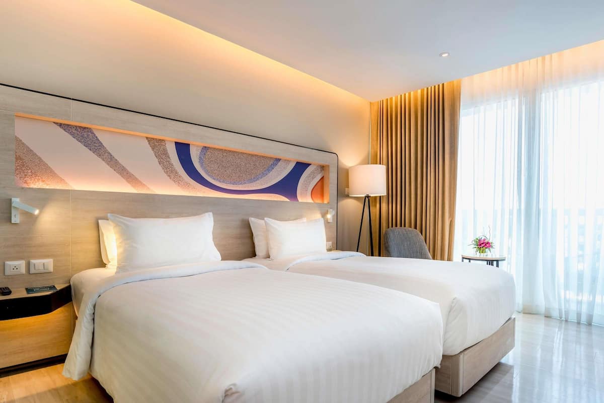 Quarto de hotel com duas camas, uma obra de arte moderna na parede, uma luminária e vista da cidade através de grandes janelas.
