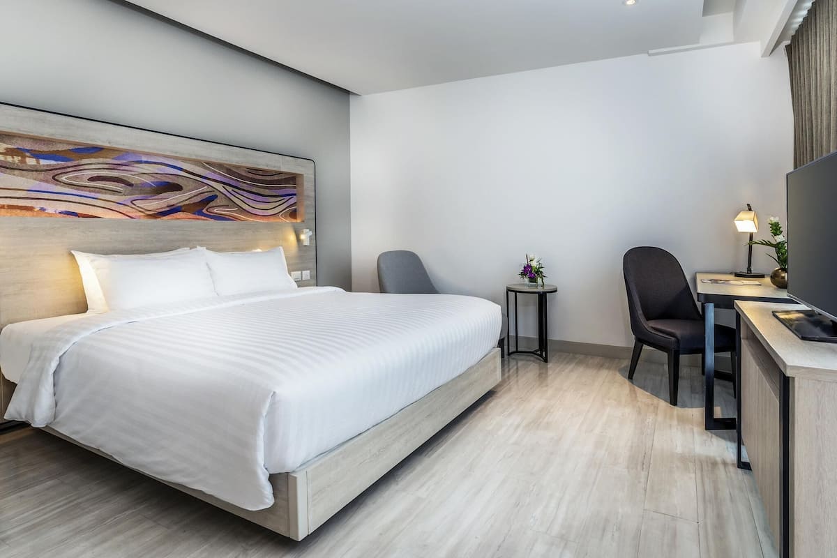 Moderni hotellihuone, jossa on suuri sänky, työpöytä tuolilla, pieni pöytä lampun kanssa ja suuri taideteos seinällä.