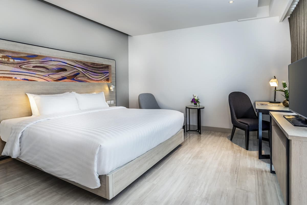 Quarto de hotel moderno com uma cama grande, uma escrivaninha com cadeira, uma mesinha de apoio com luminária e uma televisão grande.