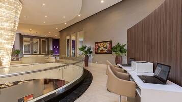 A modern lobby with a glass railing, a chandelier, a reception desk with a laptop, and potted plants.