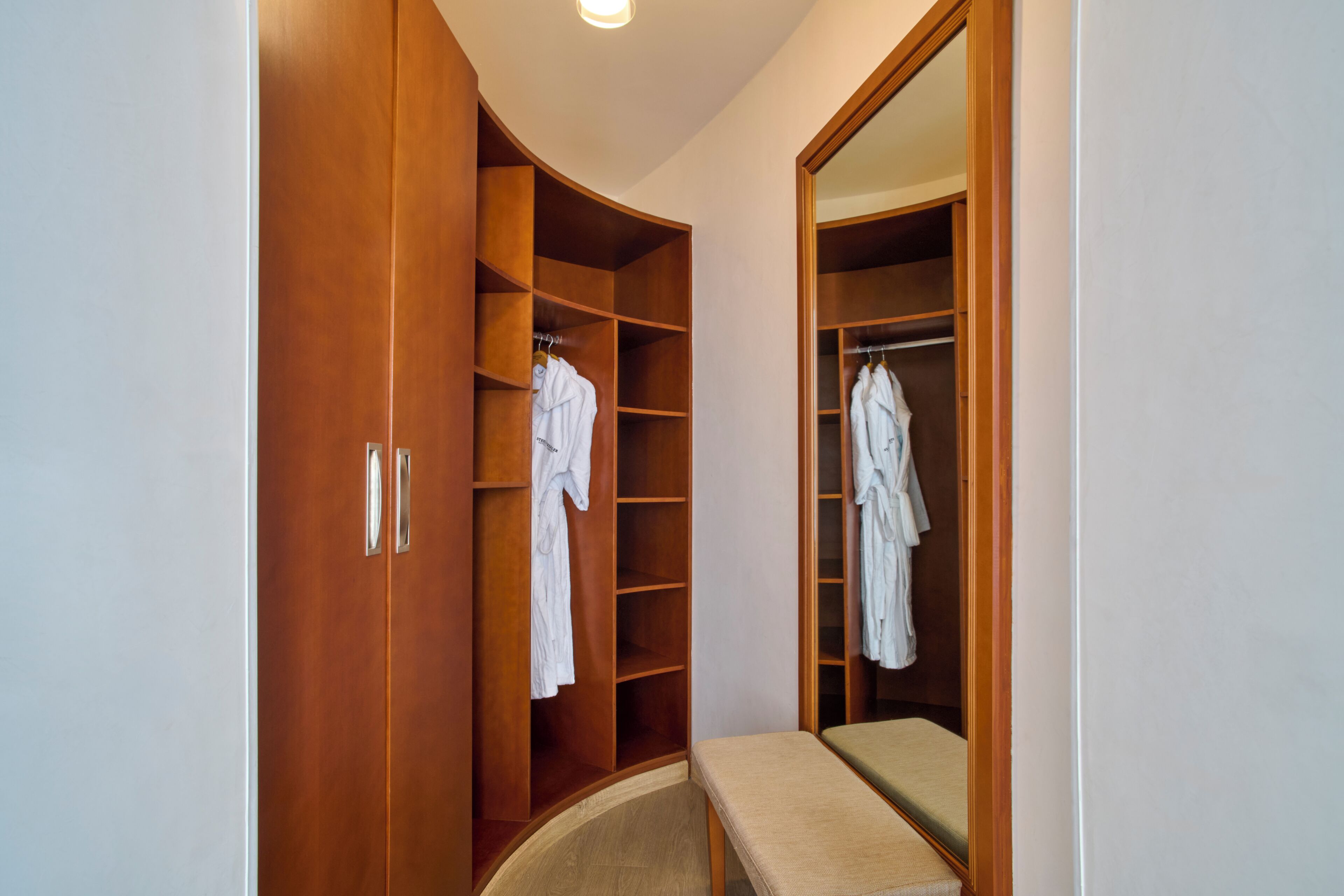 A walk-in closet with wooden doors and shelves, containing white robes and hangers.