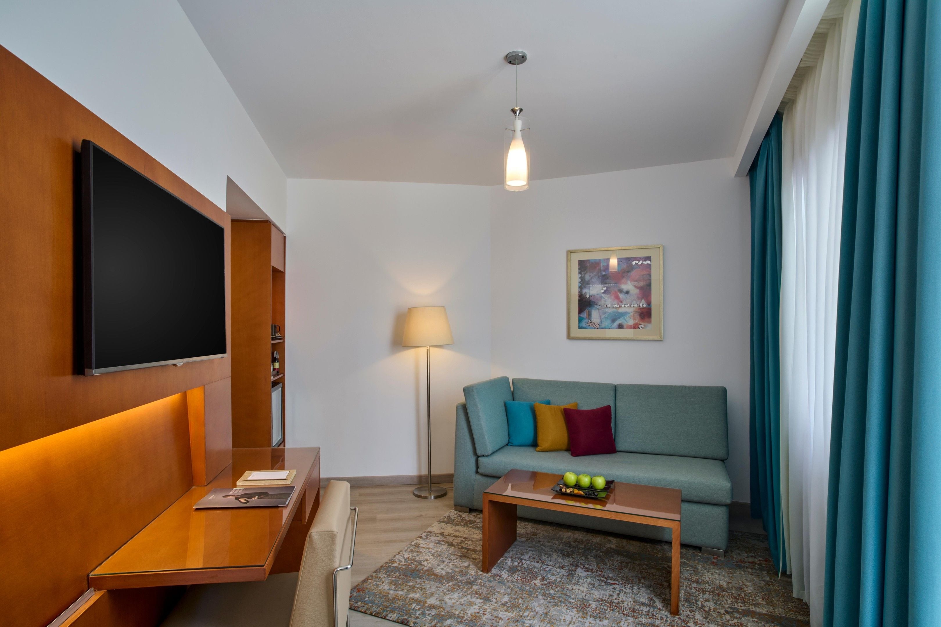 A modern hotel room with a flat-screen TV, a sofa with colorful cushions, a small coffee table with fruit, a floor lamp, and a painting on the wall.