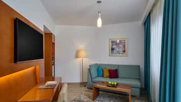 A modern hotel room with a flat-screen TV, a sofa with colorful cushions, a small coffee table with fruit, a floor lamp, and a painting on the wall.