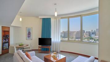 Una habitación de hotel moderna con un amplio ventanal que ofrece vistas a la ciudad, un televisor de pantalla plana, una zona de comedor y un espacio de estar con sofá y mesa de centro.