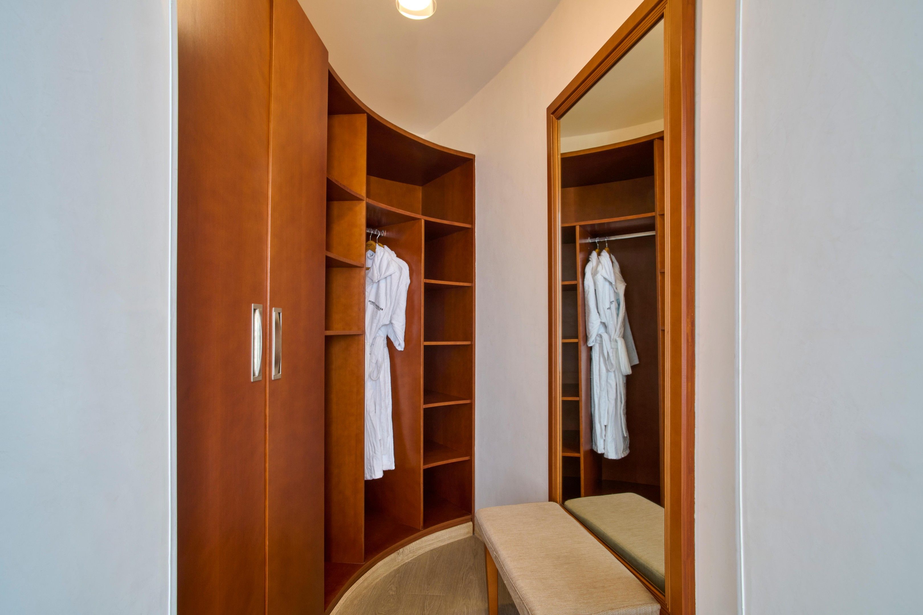 Un vestidor con puertas y estantes de madera, que contiene batas colgadas y un banco.