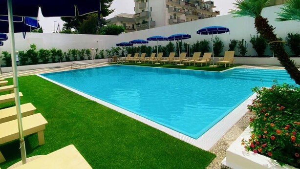 Una piscina con sillas de descanso y sombrillas, rodeada de vegetación y edificios.