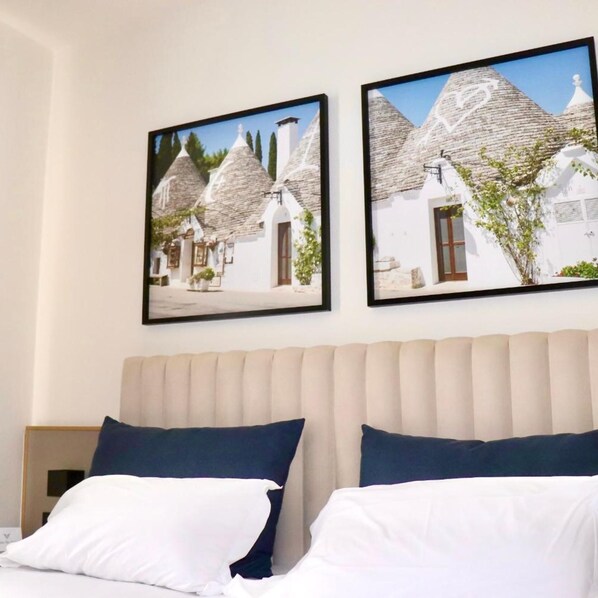 A hotel room with a bed, two framed pictures of white buildings with thatched roofs, and a plain wall.