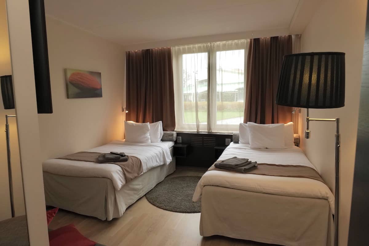 A hotel room with two single beds, a small table, a window with curtains, and a lamp.
