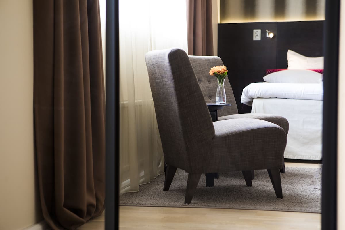 A hotel room with a bed, a chair, a small table, and a vase with flowers.