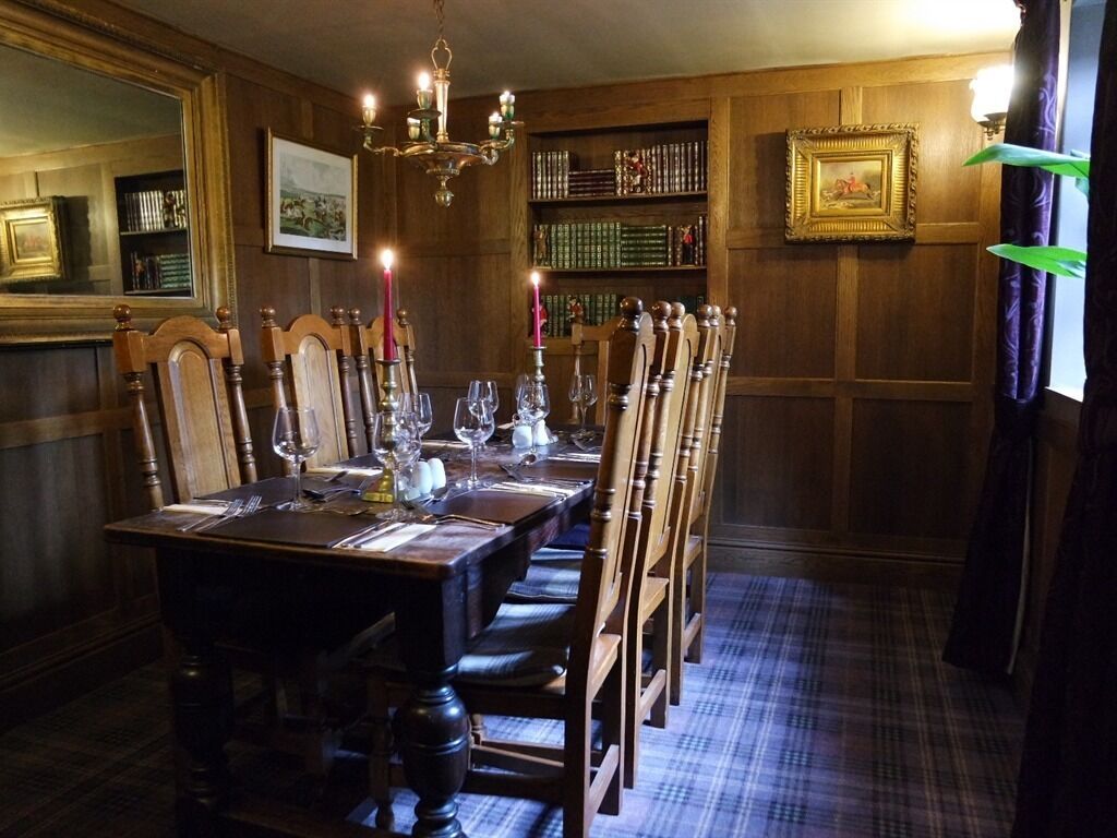 Une salle à manger avec une longue table en bois, dressée pour six personnes, entourée de chaises hautes en bois. Un lustre est suspendu au plafond, et une bibliothèque remplie de livres est visible au fond de la pièce.