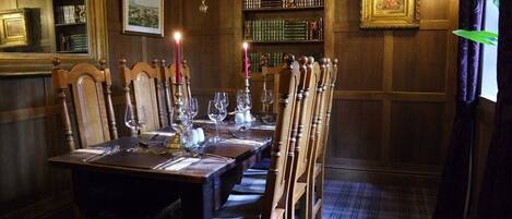 Une salle à manger avec une longue table en bois, dressée pour six personnes, entourée de chaises hautes en bois. Un lustre est suspendu au plafond, et une bibliothèque remplie de livres est visible au fond de la pièce.
