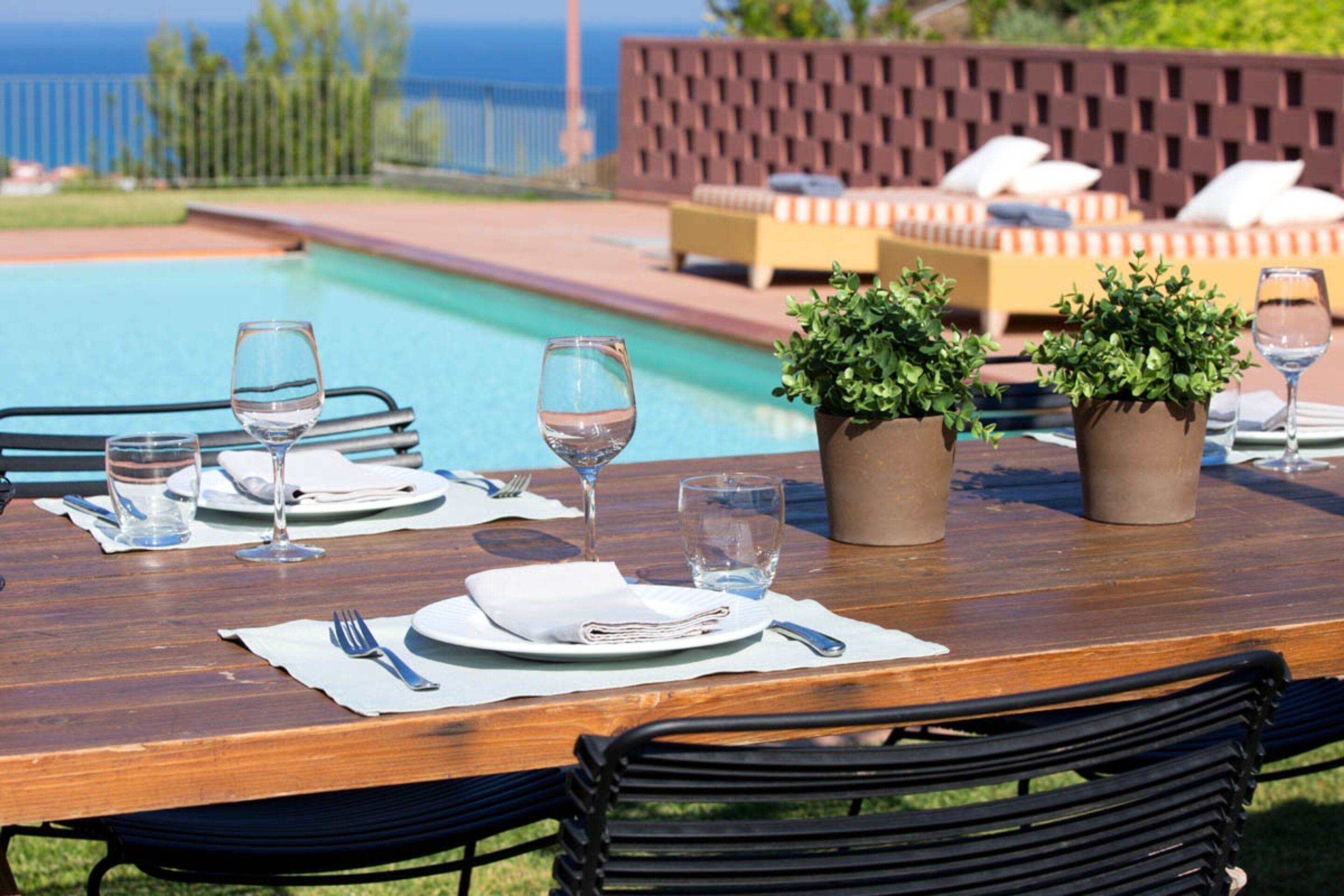 Une table en bois, dressée pour le dîner, avec vue sur la piscine, des sièges d’extérieur et des plantes en pot.