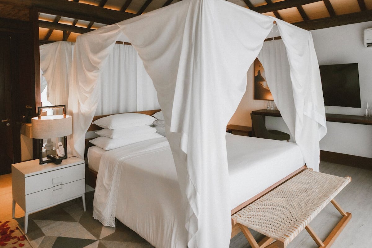 A bedroom with a canopy bed, a bedside table with a lamp, a bench, and a geometric patterned rug.