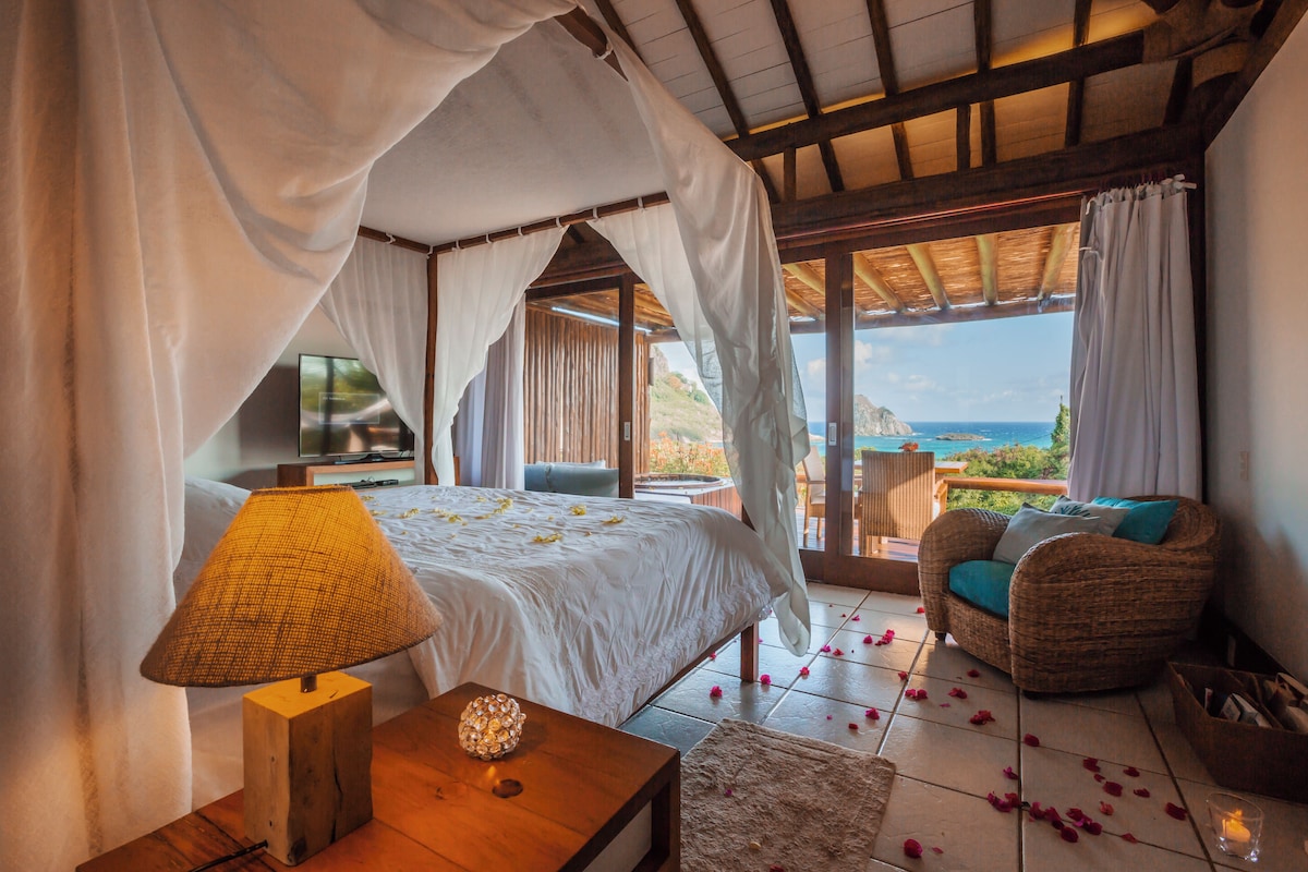 Ein Schlafzimmer mit Bett, Nachttischlampe, einem Korbstuhl und Blick auf das Meer.