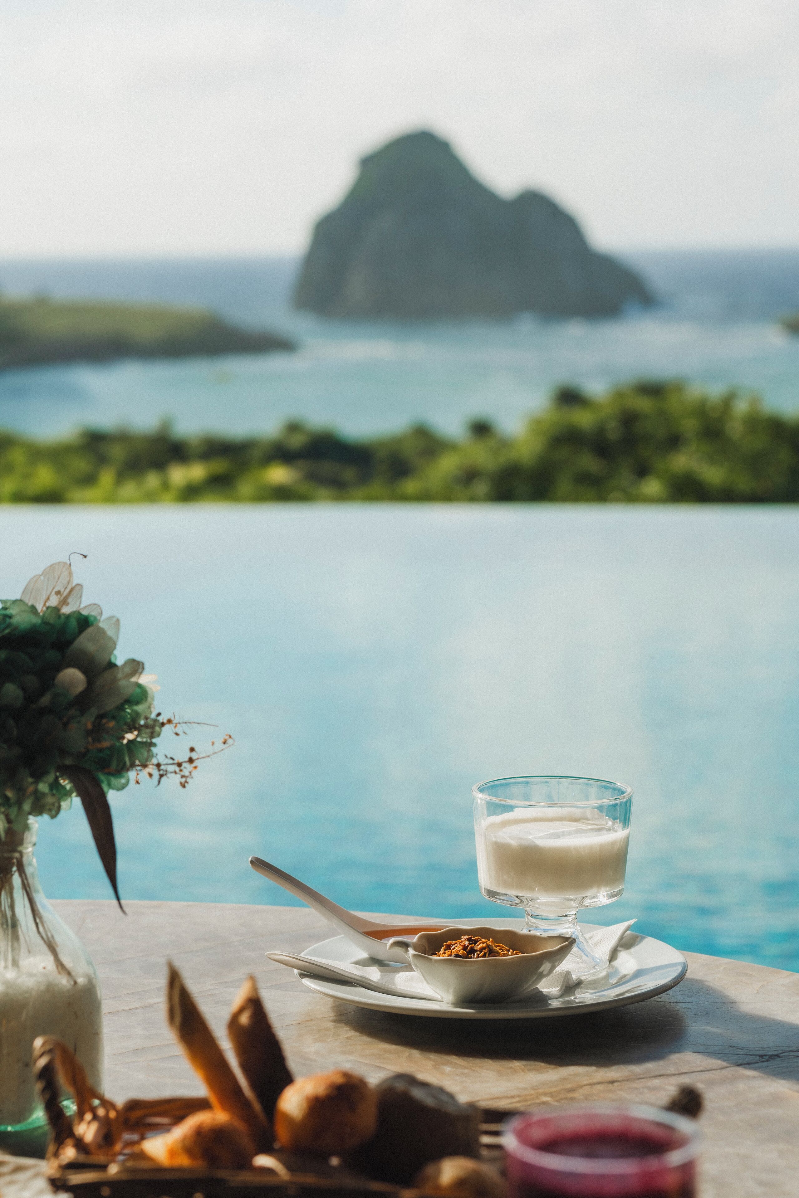 A table with a glass of milk, a bowl of granola, and a vase of flowers by a pool with a view of a mountain.