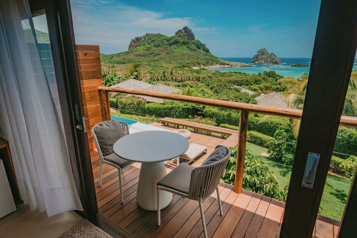 A balcony with a wooden deck, a round table, two chairs, and a view of a mountainous landscape and ocean.