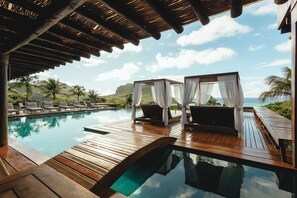 A wooden deck with a pool, lounge chairs, and a canopy-covered seating area overlooking the ocean.