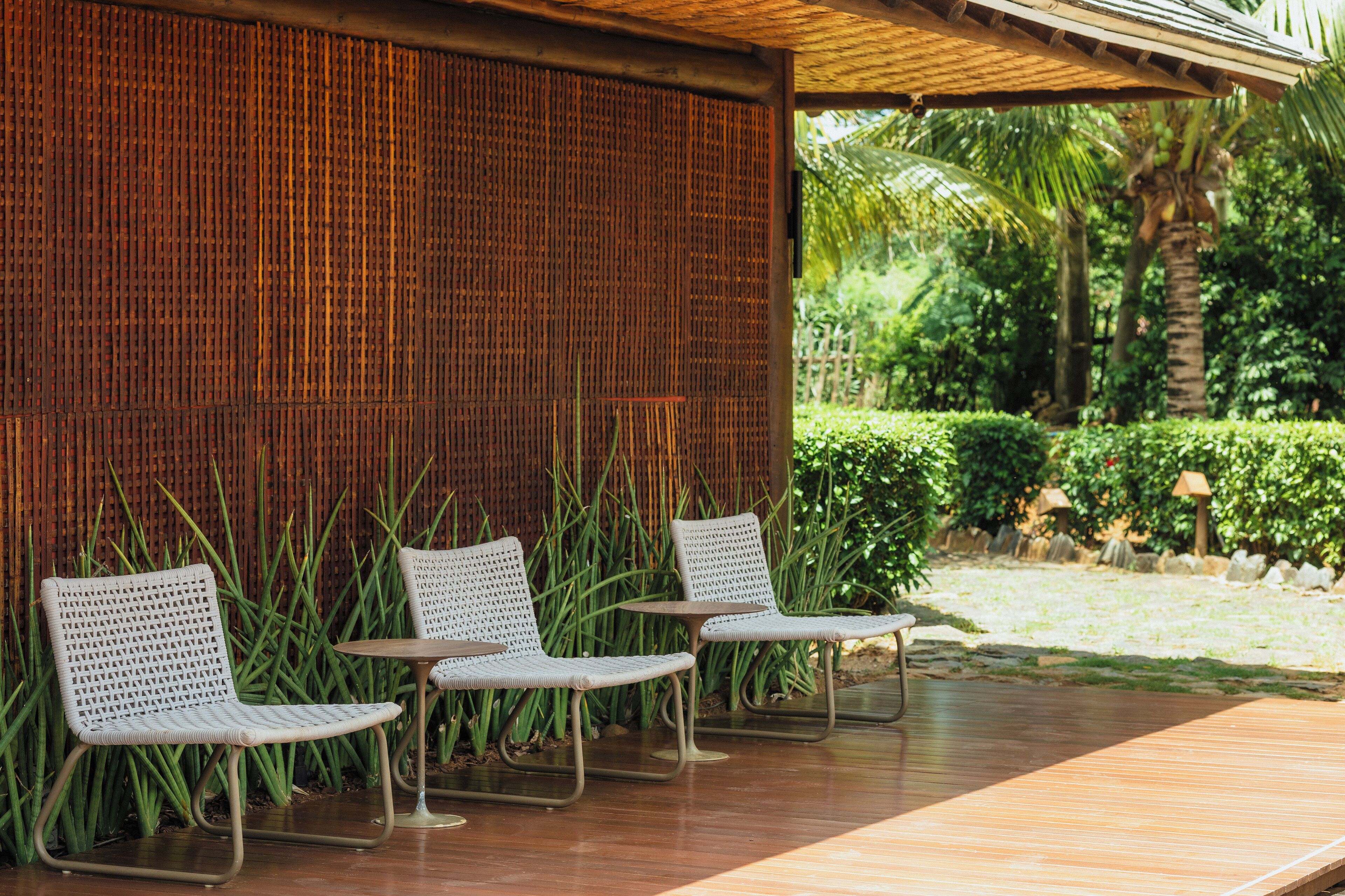 A seating area with wicker chairs and a small table, a bamboo wall, and a garden with palm trees and shrubs.