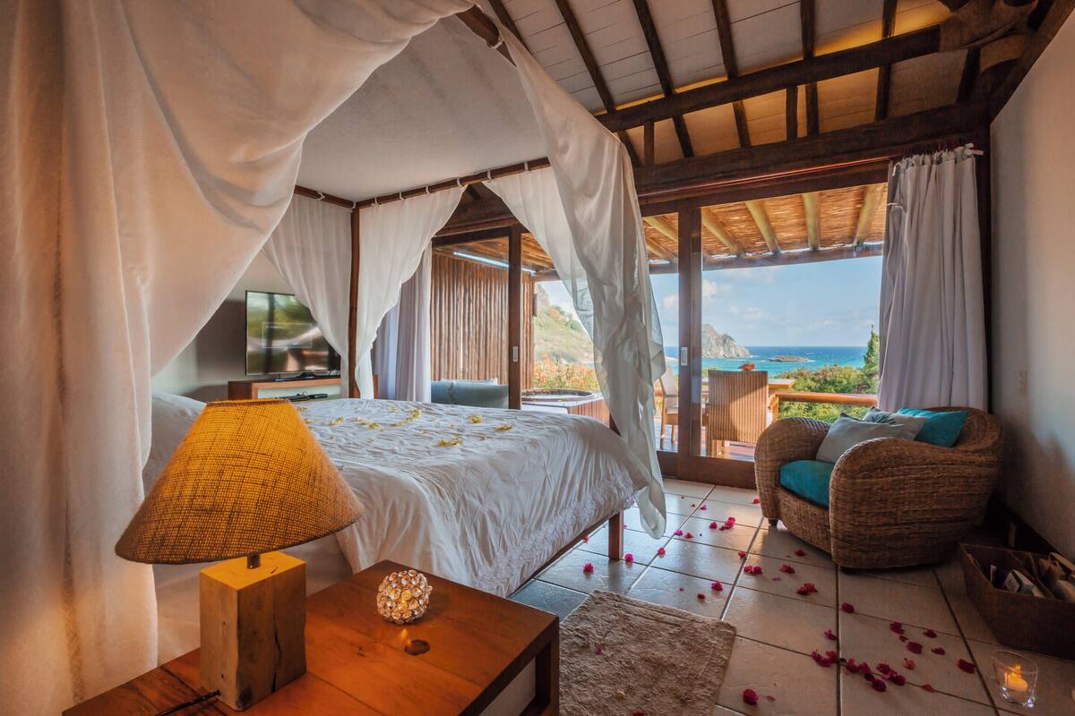A bedroom with a bed, a bedside lamp, a wicker chair, and a view of the ocean.