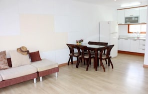 Un salon moderne comprenant un canapé, une table à manger avec des chaises et un coin cuisine doté de meubles blancs et d’appareils électroménagers.