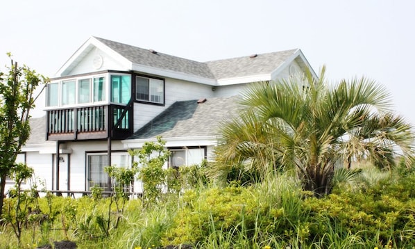 Une maison moderne à deux étages, dotée d’un balcon, et entourée d’une végétation luxuriante et de palmiers.