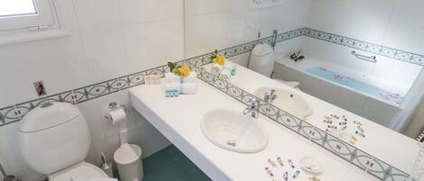 A bathroom with a white sink, mirror, and bathtub. There is a toilet with a white lid and a trash can. The floor is green, and the walls are white with a decorative border.