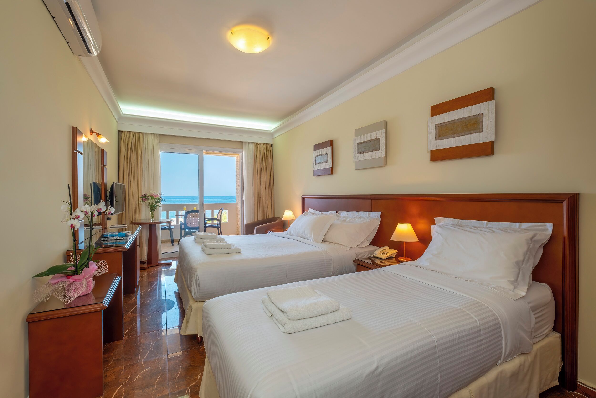 A hotel room with two beds, a desk, a chair, a lamp, and a view of the sea.
