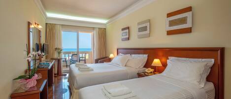 A hotel room with two beds, a desk, a chair, a lamp, and a view of the sea.