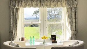 Ein Zimmer mit Blick auf ein grünes Feld und Bäume, zwei Sessel, ein Tisch mit Blumenvase und ein Fenster mit Vorhängen.