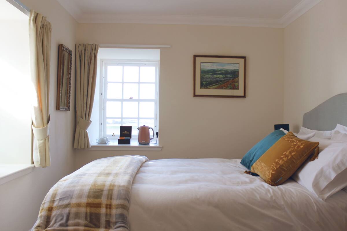A bedroom with a bed, a window with curtains, a painting on the wall, and a kettle on the windowsill.