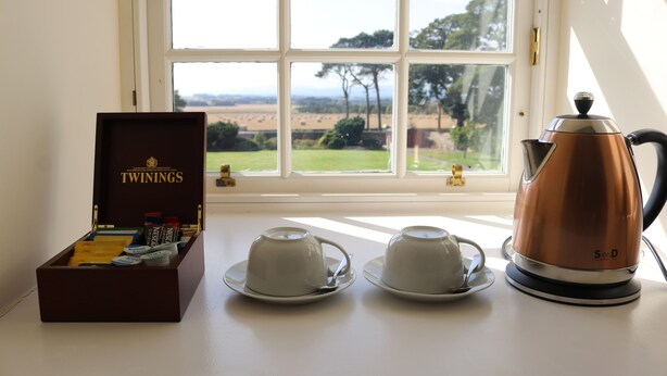 Ein Tisch mit zwei weißen Tassen und Untertassen, einer Kupfer-Teekanne und einer mit „TWININGS“ beschrifteten Schachtel mit Tee.