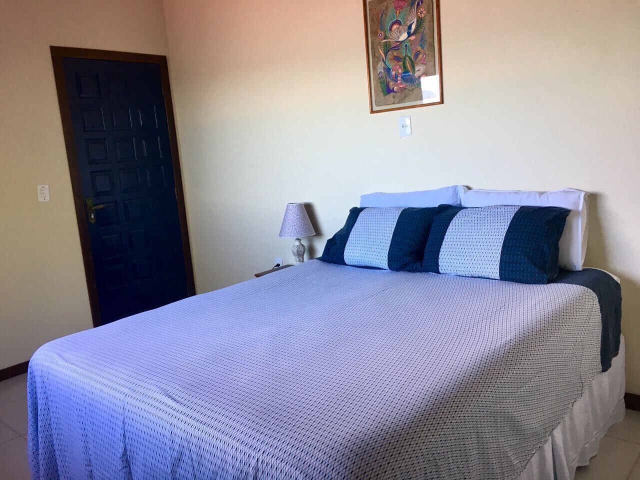 Una cama bien hecha con un edredón estampado, una mesita de noche con lámpara, y una puerta en una habitación sencilla con paredes lisas.