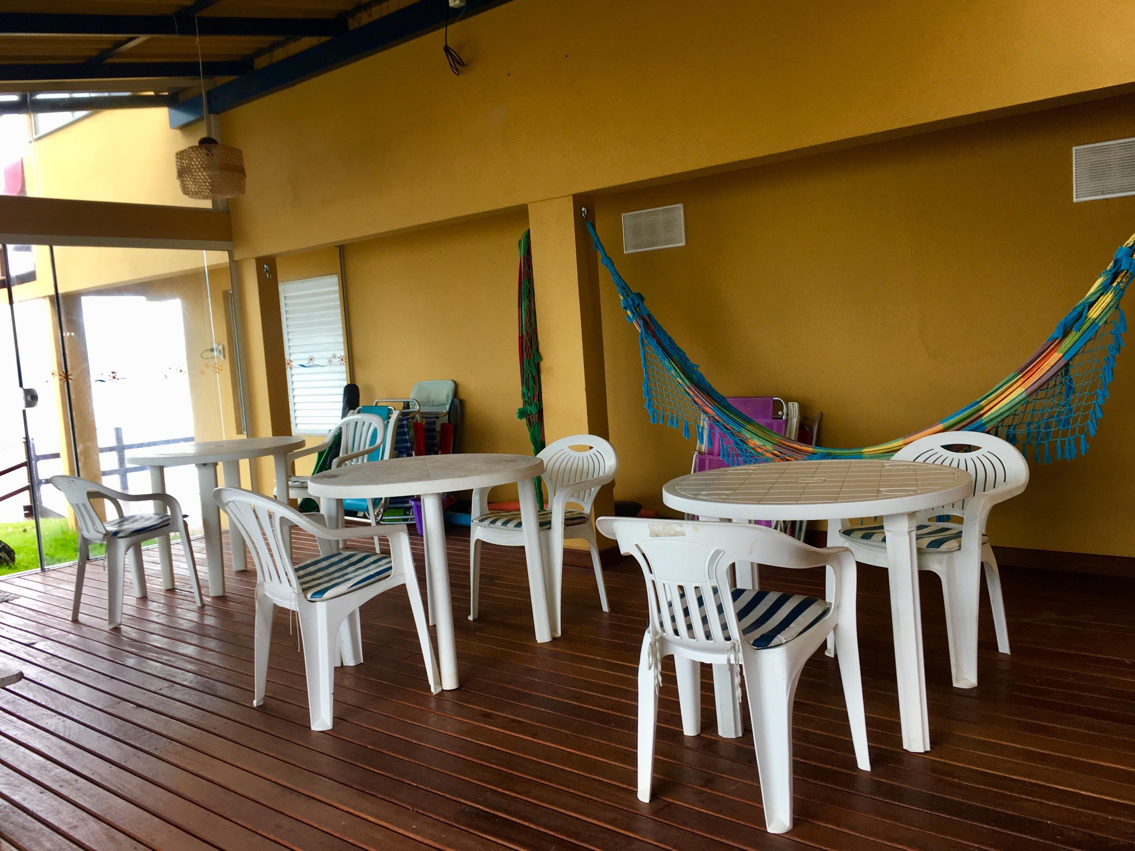 Un área de estar al aire libre con techo, con sillas y mesas de plástico blanco, una hamaca colorida y una terraza de madera.