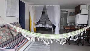 A room with a hammock, a bed with white curtains, a striped sofa, a dining table with chairs, and a kitchen area with a refrigerator and microwave. - Pousada Prainha da Nina (Florianópolis)