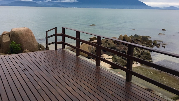 A wooden deck with a railing overlooking a calm body of water and distant mountains. - Pousada Prainha da Nina (Florianópolis)