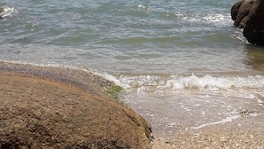 A rocky beach with pebbles and a large boulder, leading to calm sea waters under a partly cloudy sky. - Pousada Prainha da Nina (Florianópolis)
