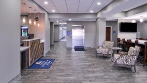 Un hall de hotel con una recepción, una zona de estar con sillas, un televisor de pantalla plana en la pared y un piso de baldosas.