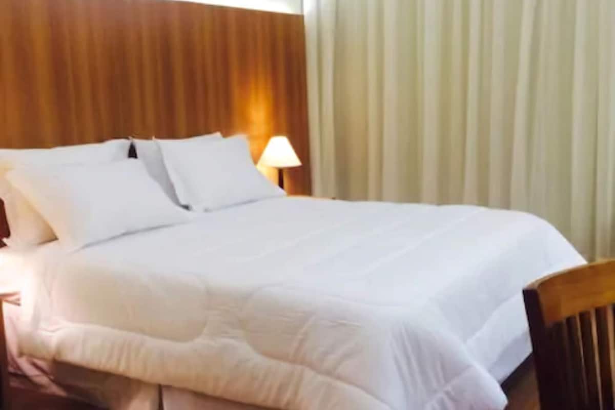 Une chambre d’hôtel avec une tête de lit en bois, un grand lit avec une literie blanche, une lampe de chevet et une fenêtre avec des rideaux.