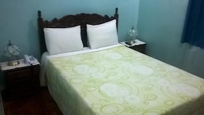 Una cama individual con colcha estampada, cabecera de madera, dos almohadas blancas, una mesita de noche con lámpara y un teléfono.