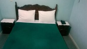 Una cama individual con cabecera de madera, almohadas blancas y colcha verde. Hay dos mesitas de noche, una con un objeto verde encima.