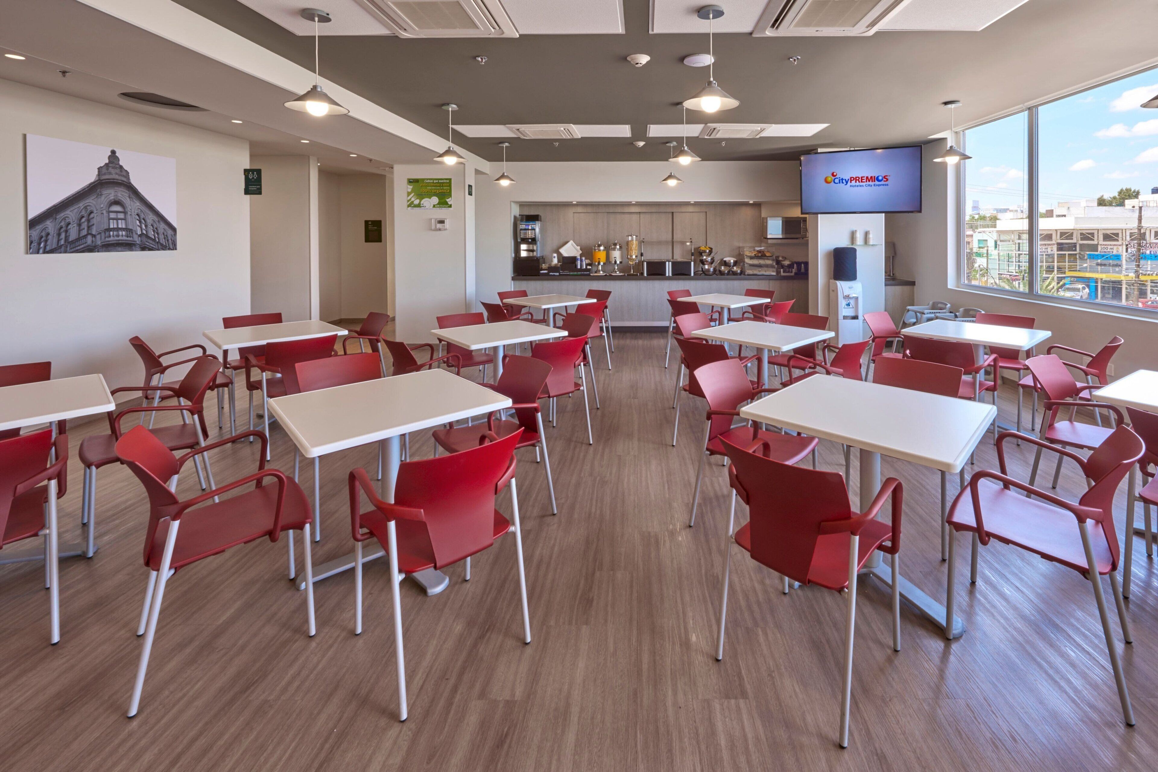 A modern dining area with red chairs and white tables, a kitchen in the background, and a large window with a city view.