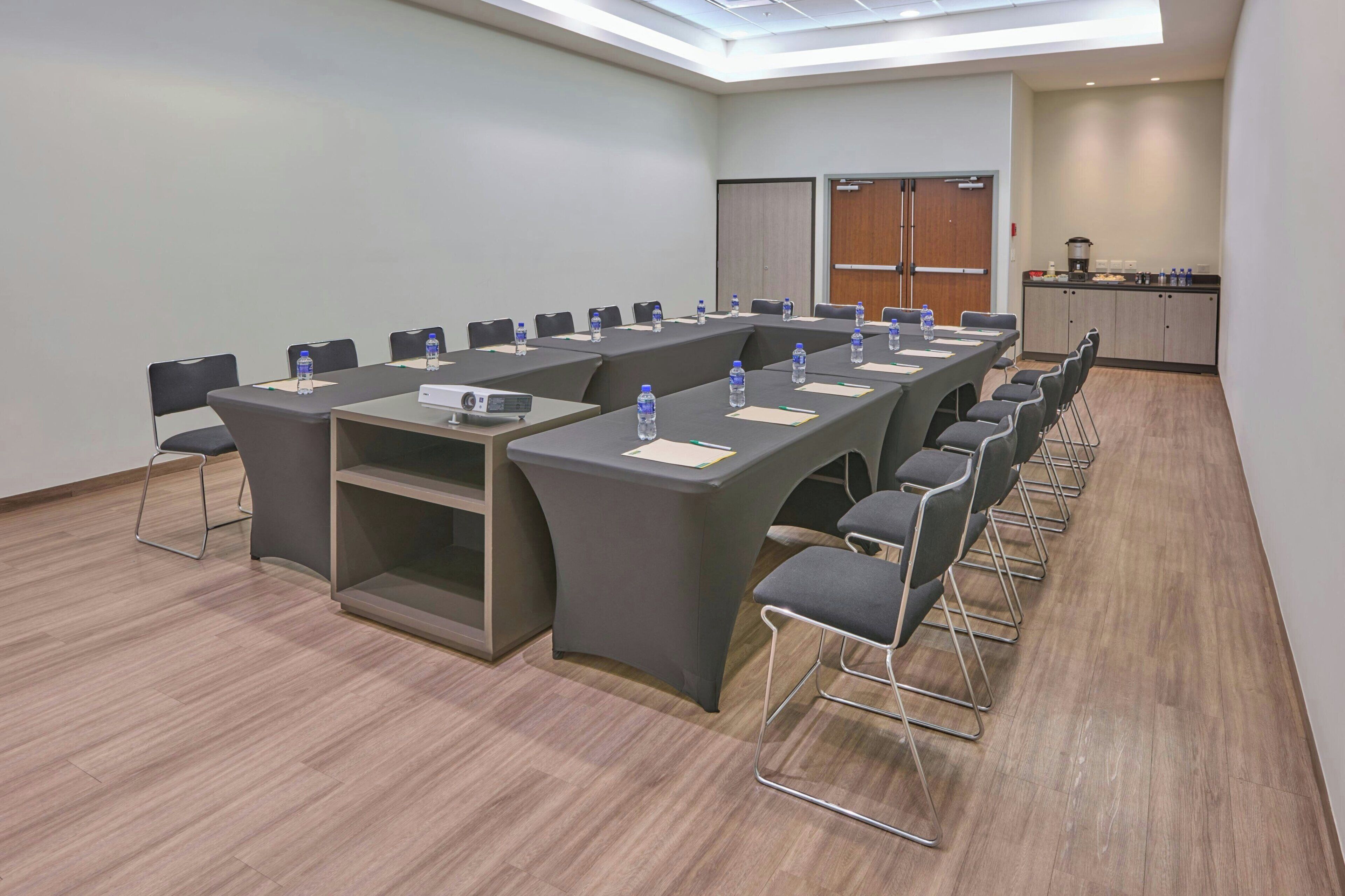 Uma sala de conferências com mesas compridas, cadeiras, garrafas de água e um pequeno espaço de cozinha.