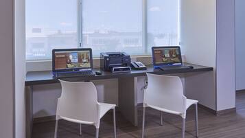Two desks with laptops and a printer, white chairs, and a large window with a view of buildings.