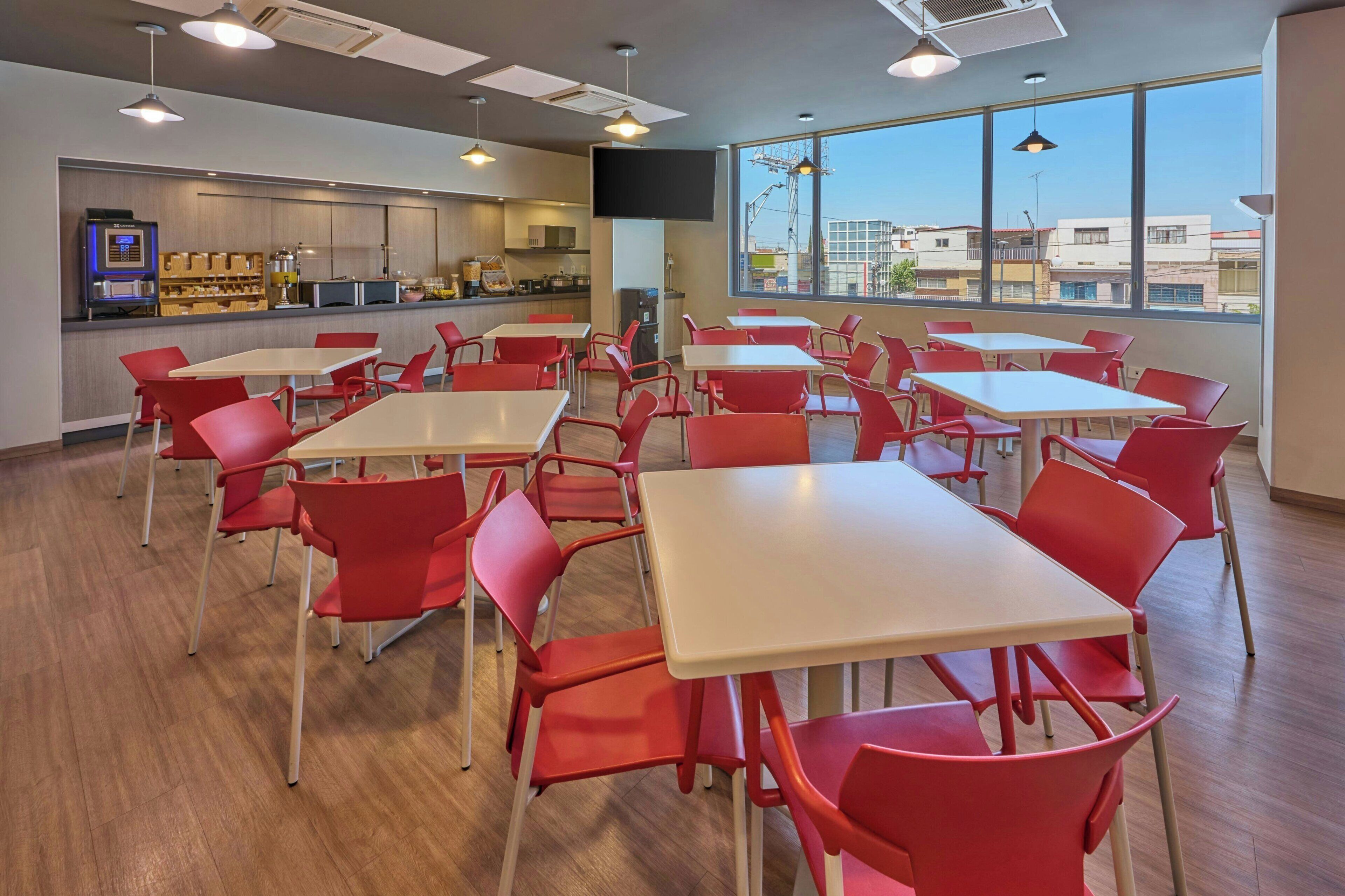 A modern dining area with red chairs and white tables, a kitchen area with a vending machine, and large windows offering a city view.