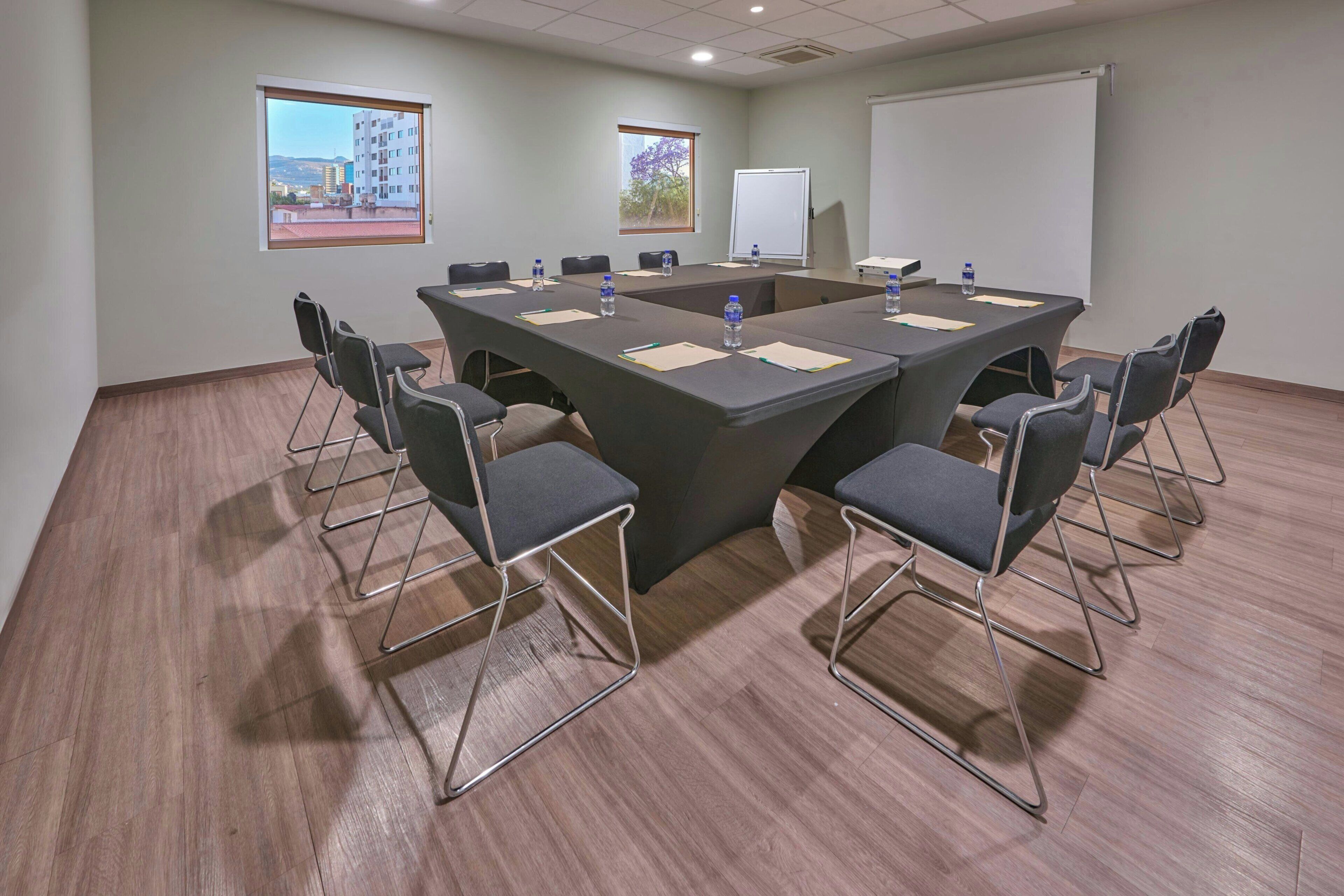 A conference room with a large table, chairs, a projector screen, and windows with views of buildings.
