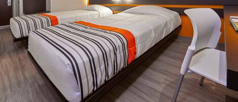 A hotel room with two beds, a white chair, and a wooden desk.