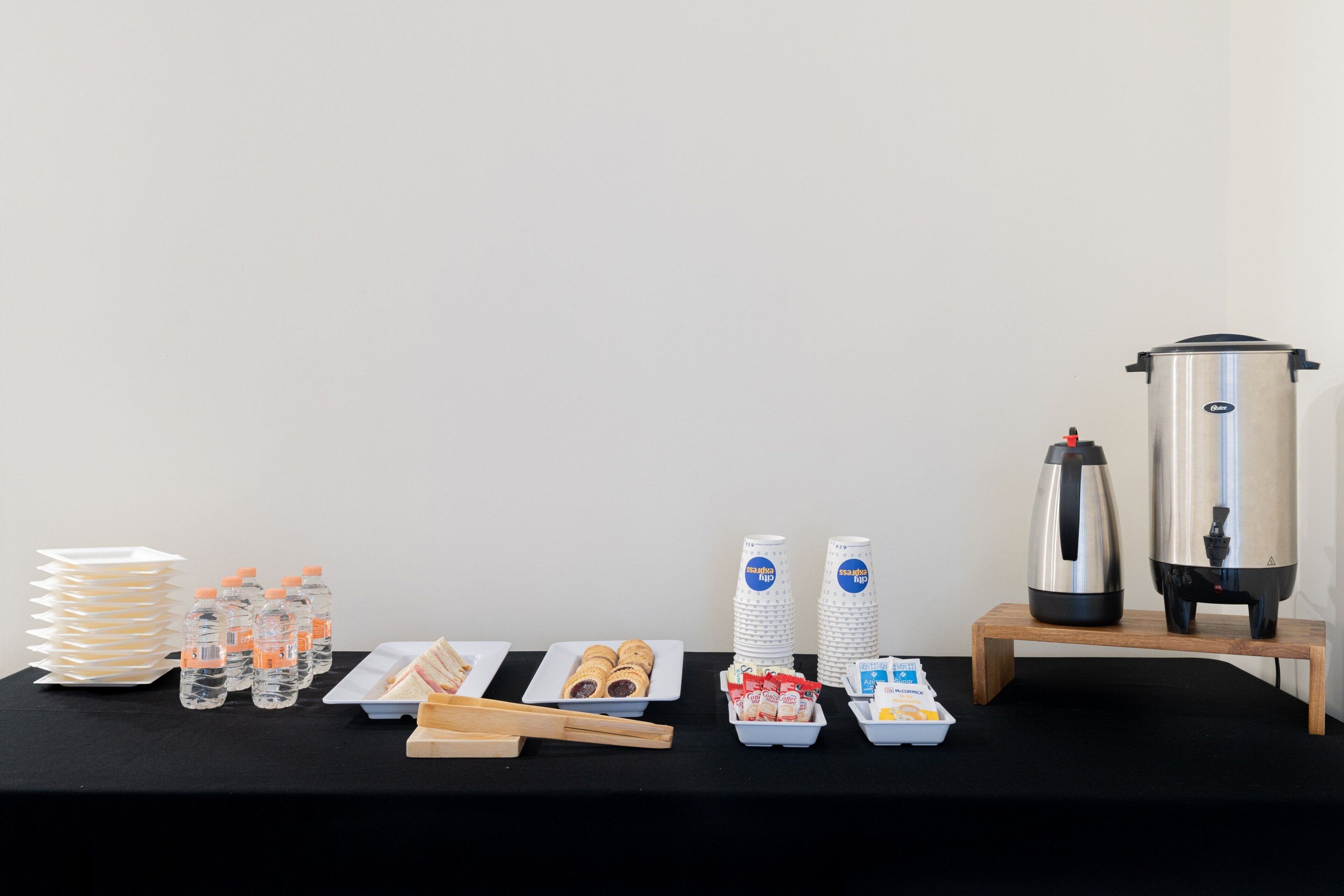 Una mesa con botellas de agua, vasos desechables, una cafetera y alimentos.
